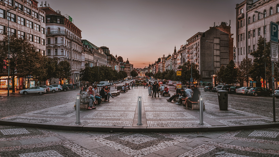Sunset view of a bustling city street with people sitting on benches and Reliance Foundry R-6321 Elysium lit bollards installed along the walkway for pedestrian safety.
