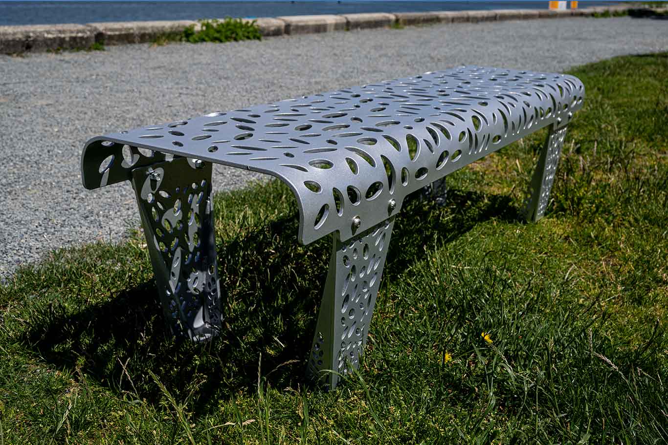 A silver bench sits on the grass beside a seawall, decorated with laser cut organic designs