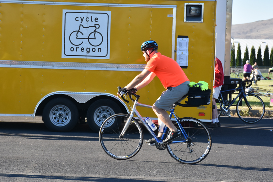 A yellow truck with the words Cycle Oregon is parked on a curb; a cyclist rides by it.