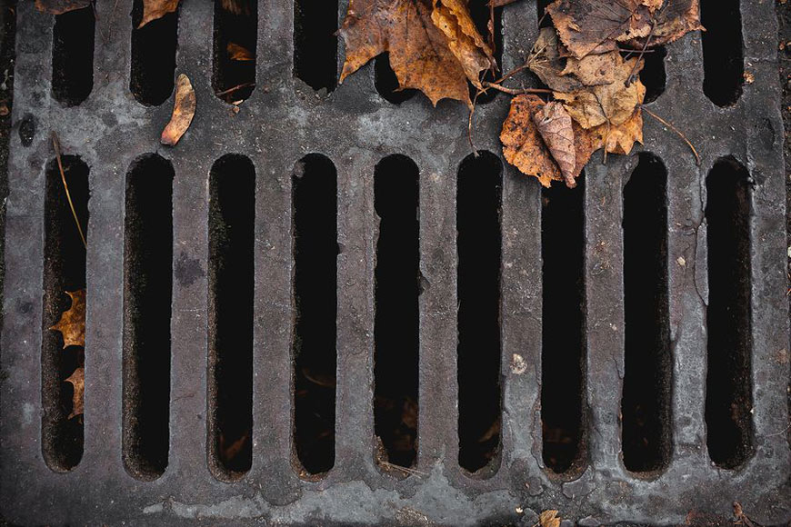 A grey iron storm sewer has developed a patina, is covered in leaves