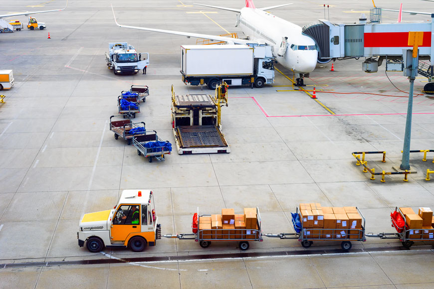 An airplane is pulled up to a gate while airport trucks move across hardscape with airport trench grates