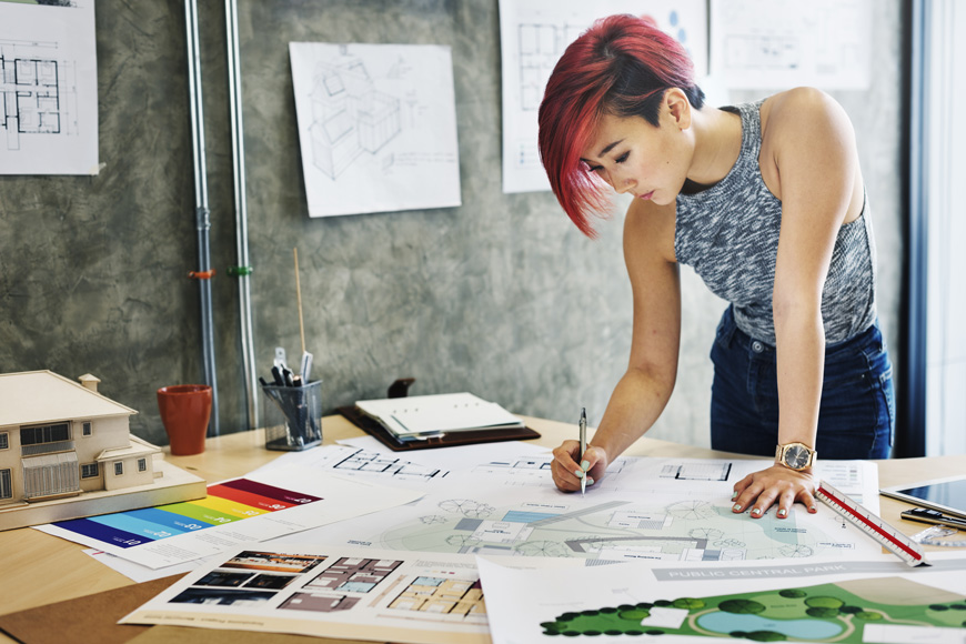 A pink-haired young woman leans over architectural and landscape drawings at standing desk
