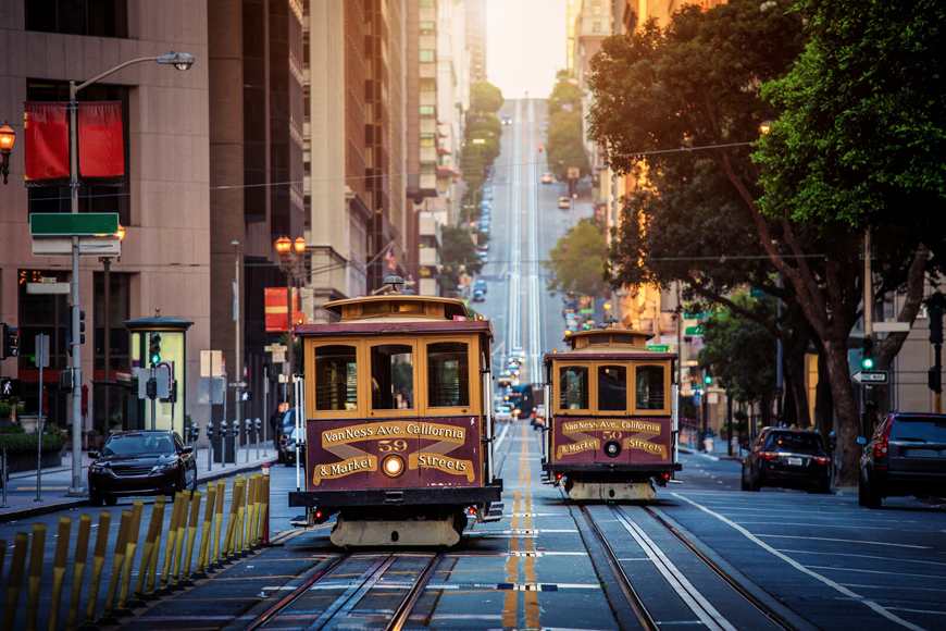 Two San Francisco trolleys with red and gold paint travel along a street at sunrise