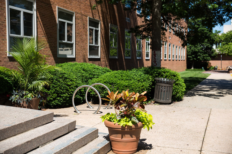 Two circle bike racks in stainless steel are affixed outside a red brick building surrounded by bushes.