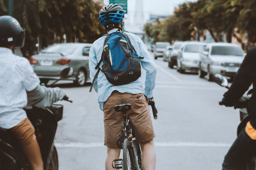 A man riding a bike waits at a light wearing a backpack and short-sleeve blue oxford shirt.
