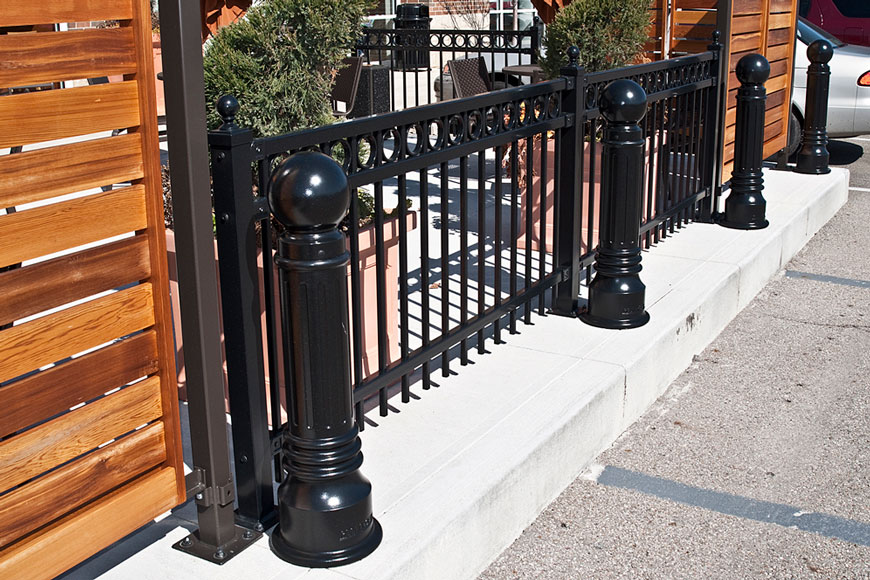 Two black, classical bollards with spherical caps protect a wrought iron fence.