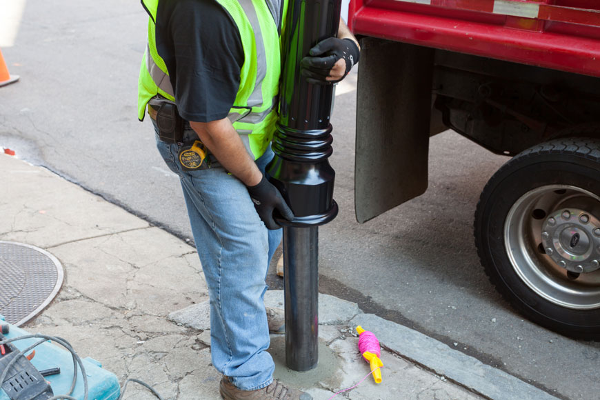 A worker wearing jeans and a high visibility vest slides a black ornate bollard over a steel and concrete security pole