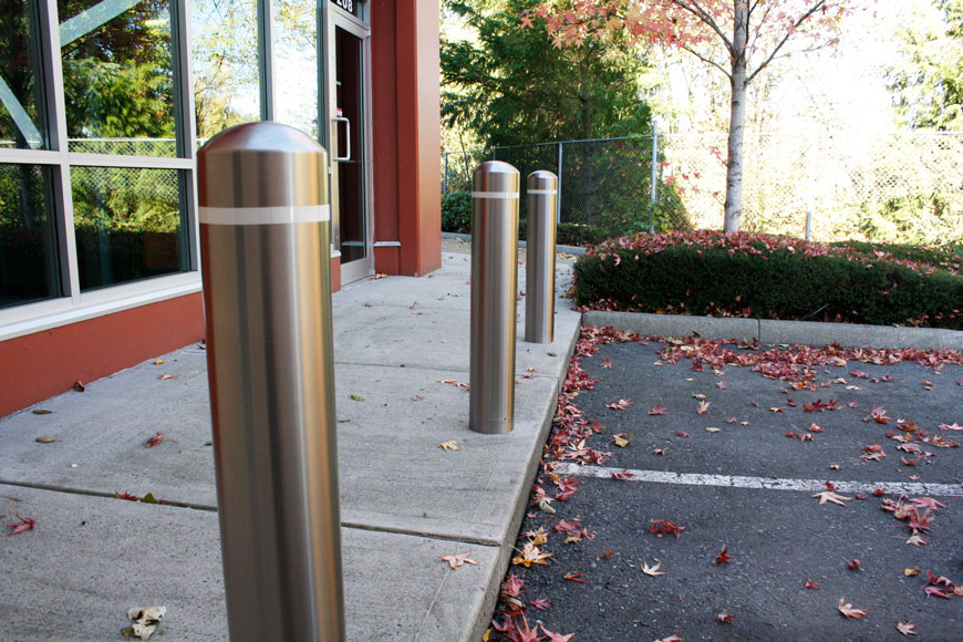 Stainless steel bollards sit on the curb above a leaf-strewn parking lot in front of a storefront