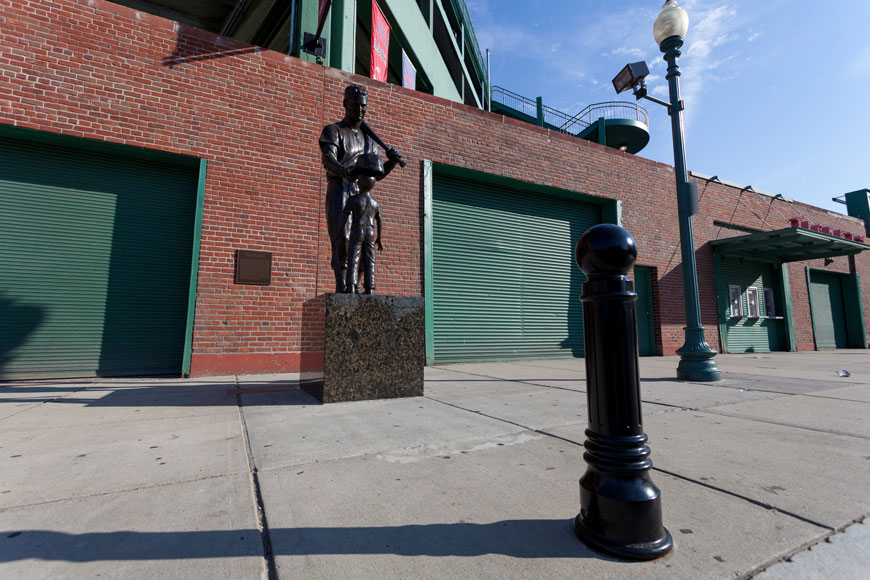 A bollard sits by a green lamp post and the Ted Williams statue of a ball player and a child fan at Fenway.
