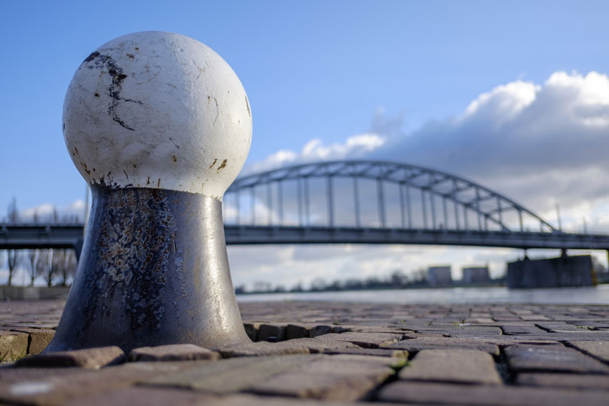 A bulbous marine bollard sits in the foreground with a bridge behind and above it