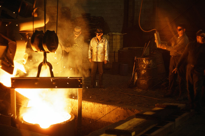 A group of foundry workers wearing goggles stand around a ladle full of molten metal