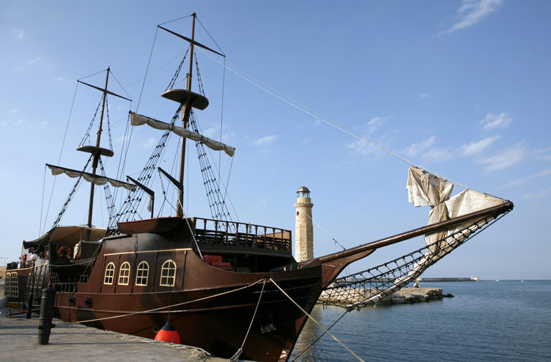 A brown “pirate-ship” with masts for canvas sails, topped with crows’ nests, tied up by a dock