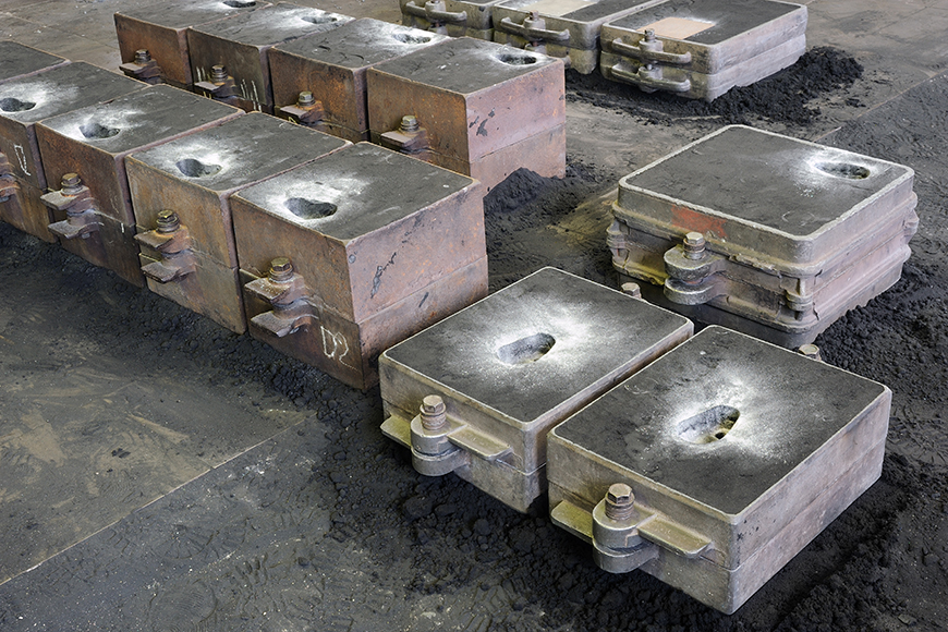 Three lines of variably sized casting flasks sit on a dirt foundry floor while their castings cool