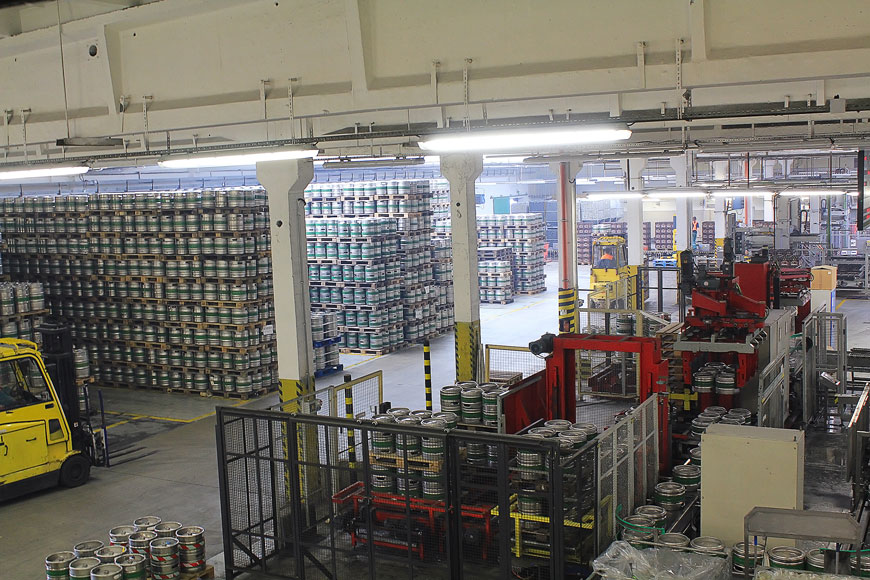 A warehouse full of stacked palettes of beer of kegs, with forklifts and industrial lighting