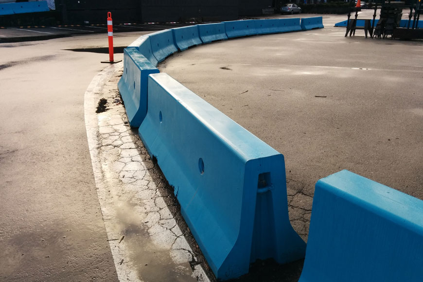 A curving line of blue concrete barriers protect a paved area from vehicle traffic