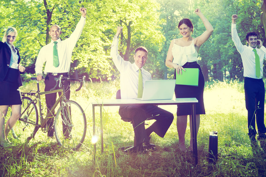 A group of bicycling advocates in business wear sit outside in a park, one standing beside a bike