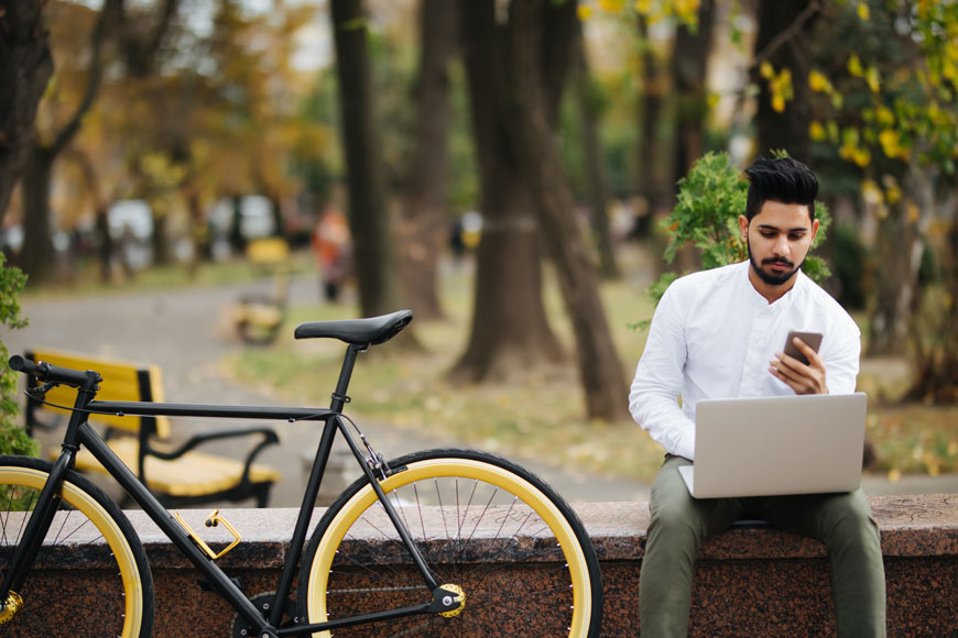 A cyclist sits in a park next to his bike, checking social media for cycling news on his phone and laptop.