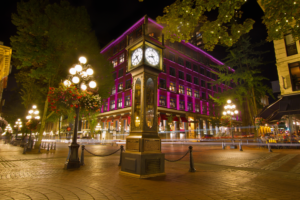 A glowing night scene features a vintage steam clock on a cobblestone street with a purple-lit building behind it