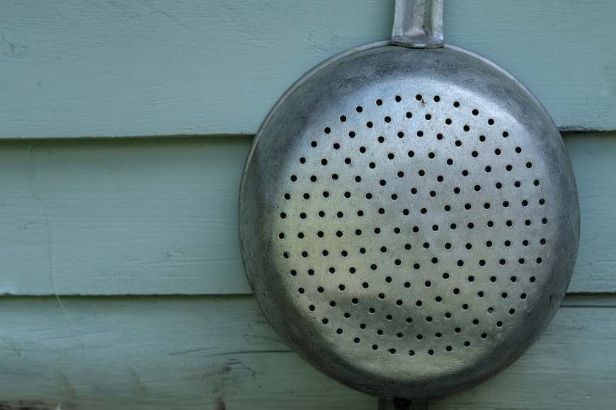 A dented aluminum colander hangs against a green painted clapboard wall.