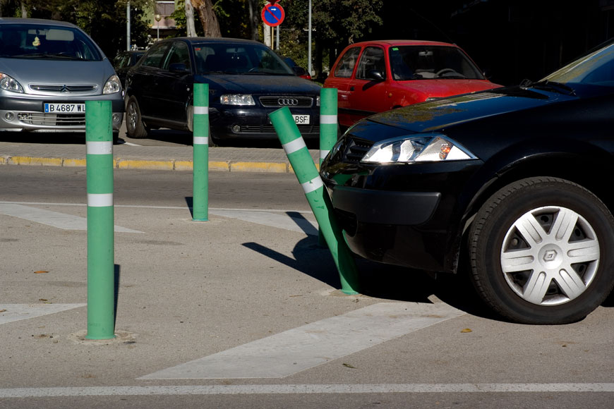 A green bollard bends as it is nudged by a black car in a parking lot
