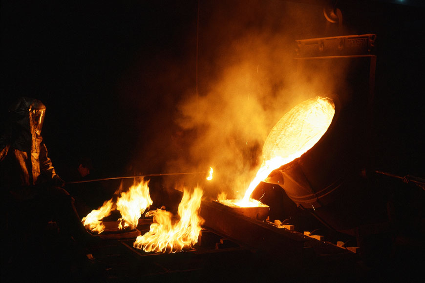 A foundry worker in a silver heat suit ladles flaming molten metal from a crucible