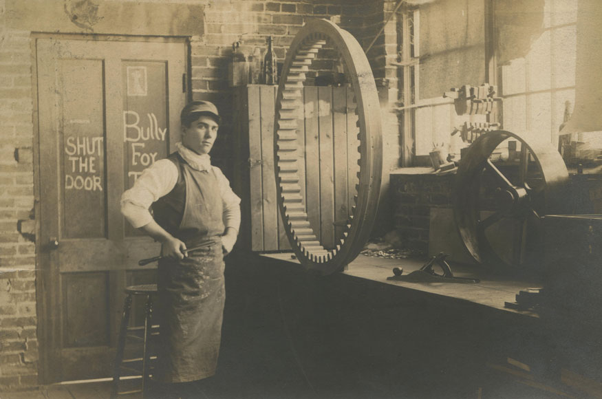 A sepia toned image from a vintage postcard shows a young man in front of a large wooden gear