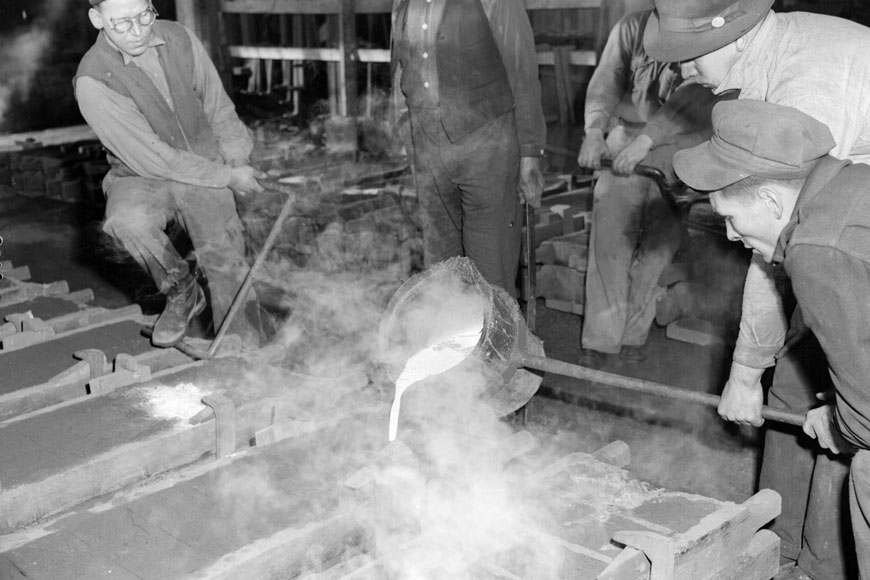 Foundrymen pour molten iron into a sand casting mold in an old black and white photograph