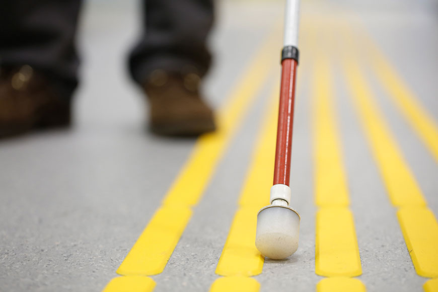 A person uses a white cane on yellow strips of tactical ground surface indicators