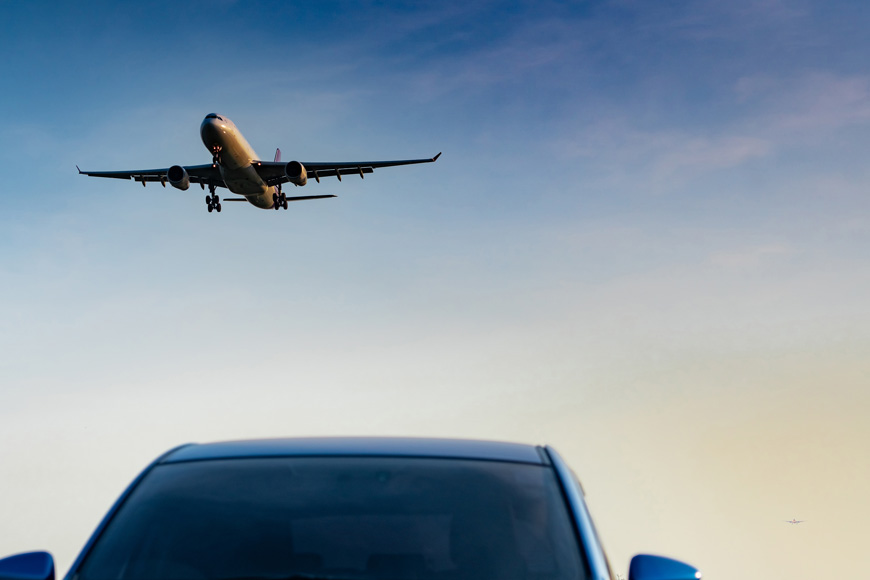 A plane takes off above the windshield of a blue car parked at an airport