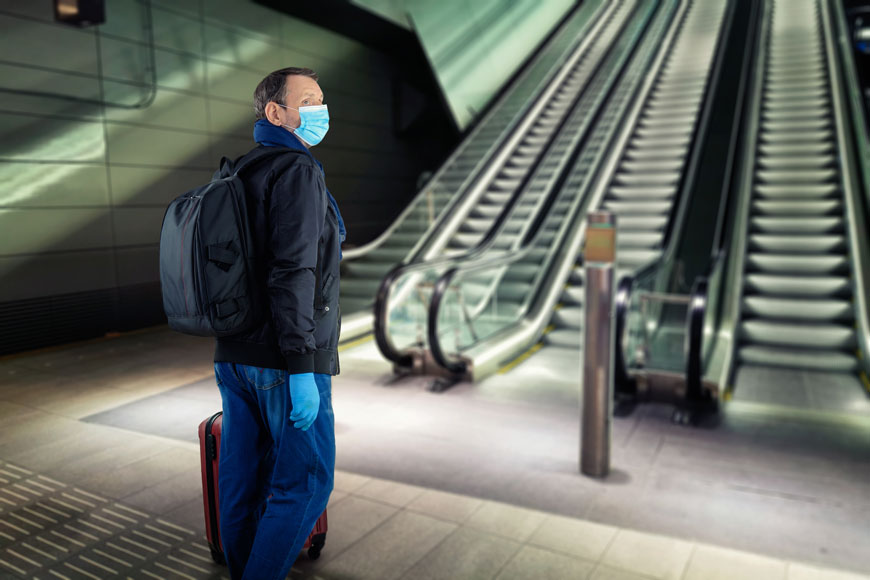 A man in a blue paper surgical mask stands near an escalator with a bollard before it.