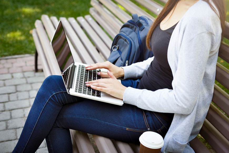 A woman sits on a bench with a laptop, her coffee beside her