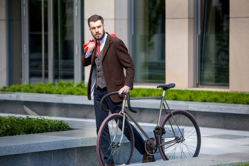 A bearded man in his 20s wearing an old-fashioned suit with vest walks beside his fixie bike.