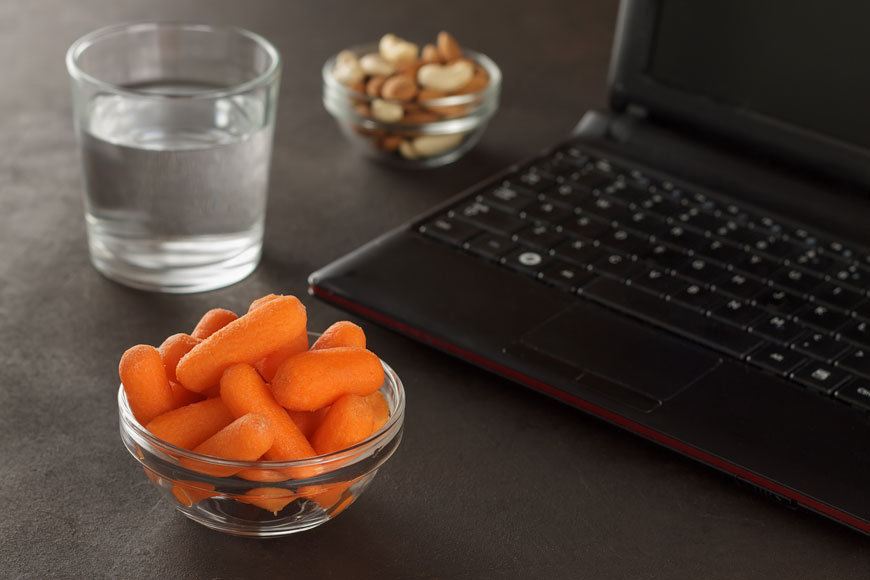 Close-up of a laptop, a bowl of baby carrots, a bowl of cashews, and a glass of water