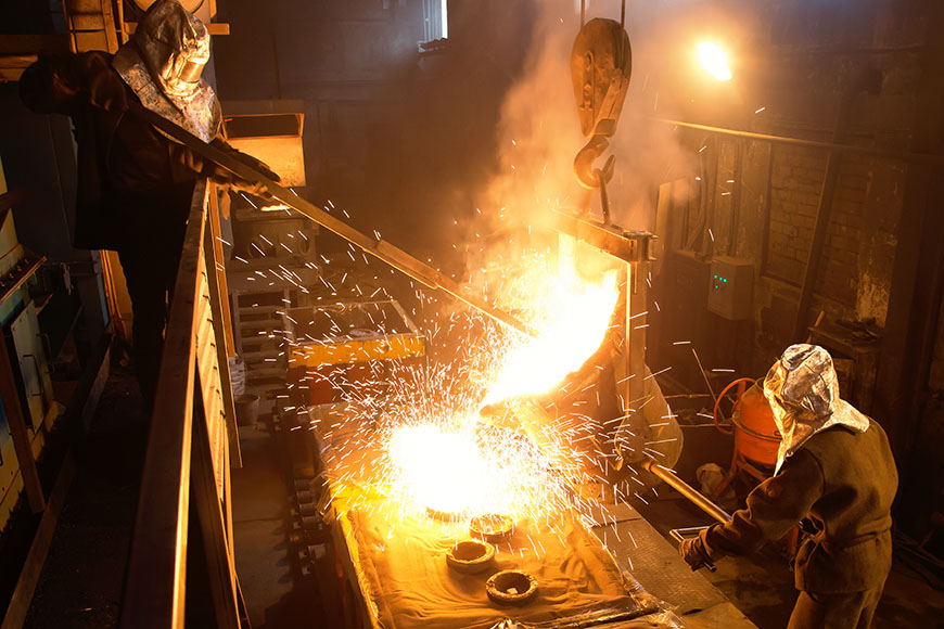 Two foundry workers in silver heat protective outfits pour metal into a mold using a tilting ladle