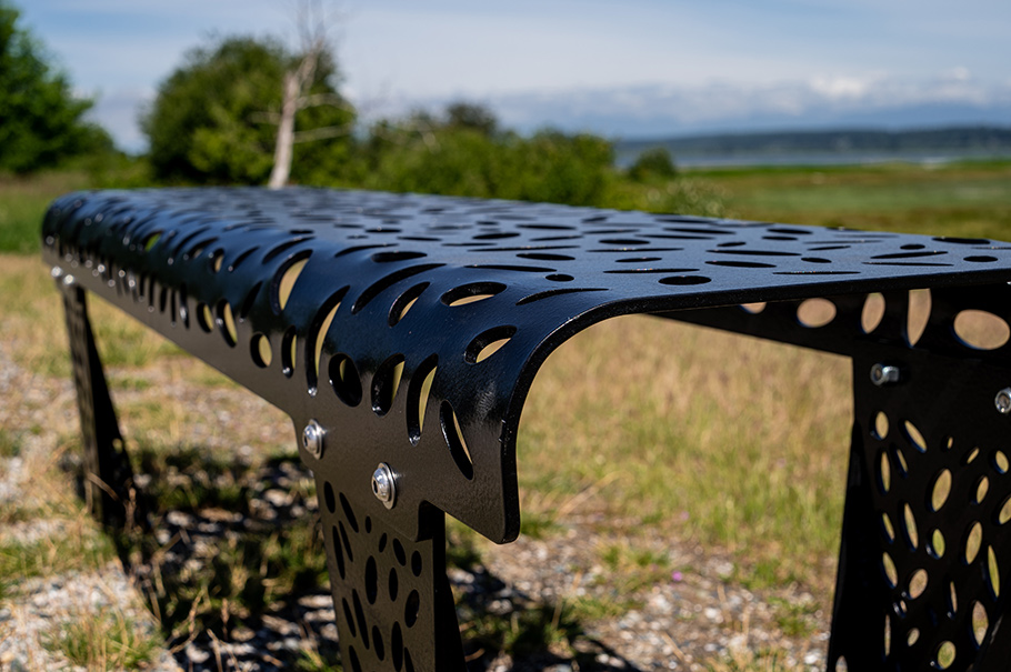 A silver bench cut with lacy designs sits by a seawall walk