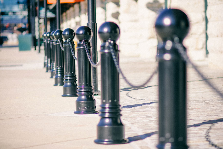 A line of black bollards that look like chess pawns stand linked by black chain near a stone building