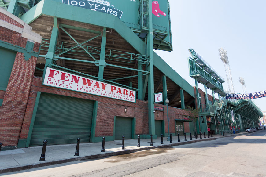 A stretch of sidewalk beside Fenway Parks' Gate C shows the green painted iron of the stands and the red-brick of the building