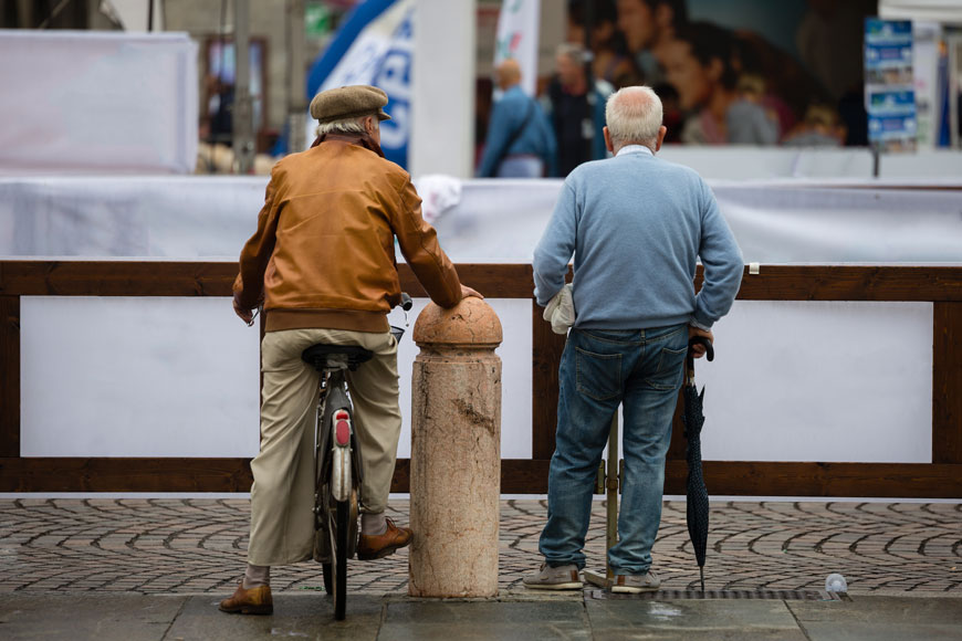 Elderly friends, one on a bicycle, stand beside protective bollard in front of low wall