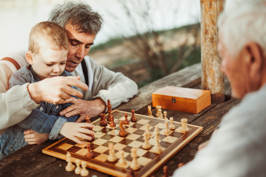Two older men play chess in a park: one holds his grandson on his knee.
