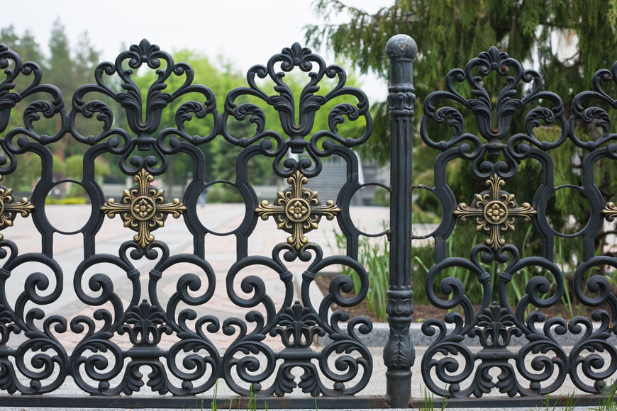A decorative ancient black filigree fence with brass floral accents is beginning to rust at joins