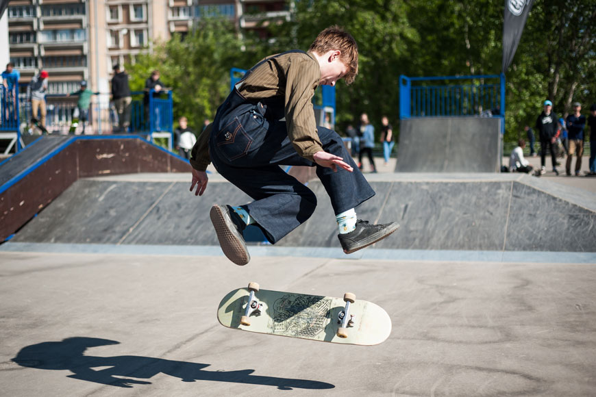 A boy wearing blue overalls and a striped shirt is caught mid-trick, hovering in the air above his turning board