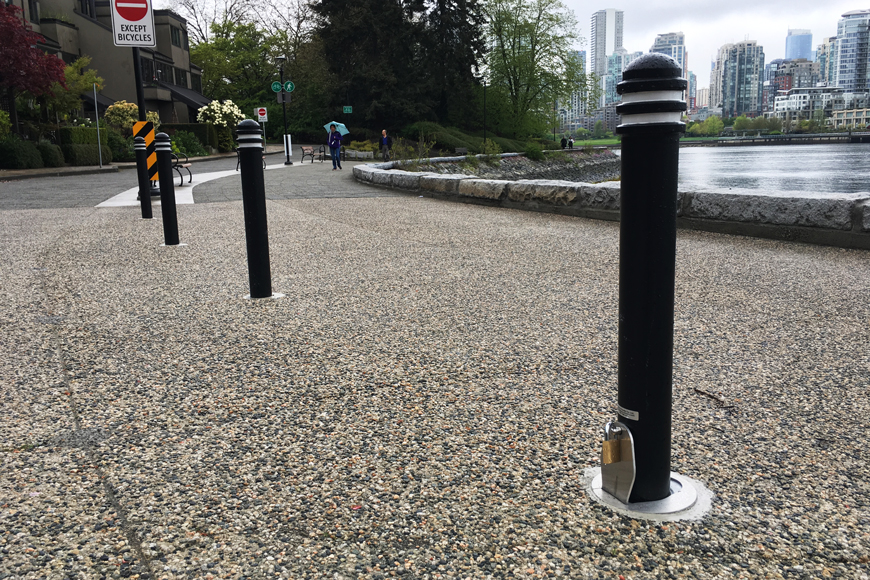 A line of bollards, one sporting a padlock, separates foot and bike traffic on a rainy seaside path.