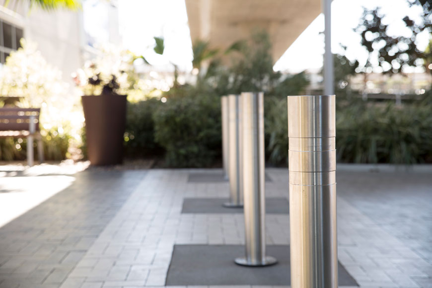 Brushed stainless steel bollards sit in a brick-laid path. Out of focus greenery in background.