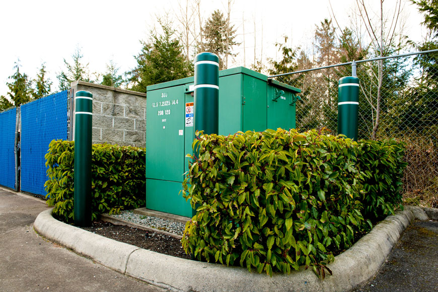 Three large security bollards covered in plastic sleeves protect a utilities box.