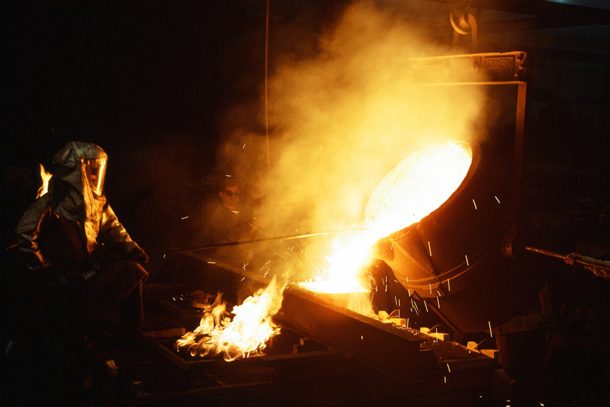 A man dressed in a silver protective suit pours red-hot metal into a mold