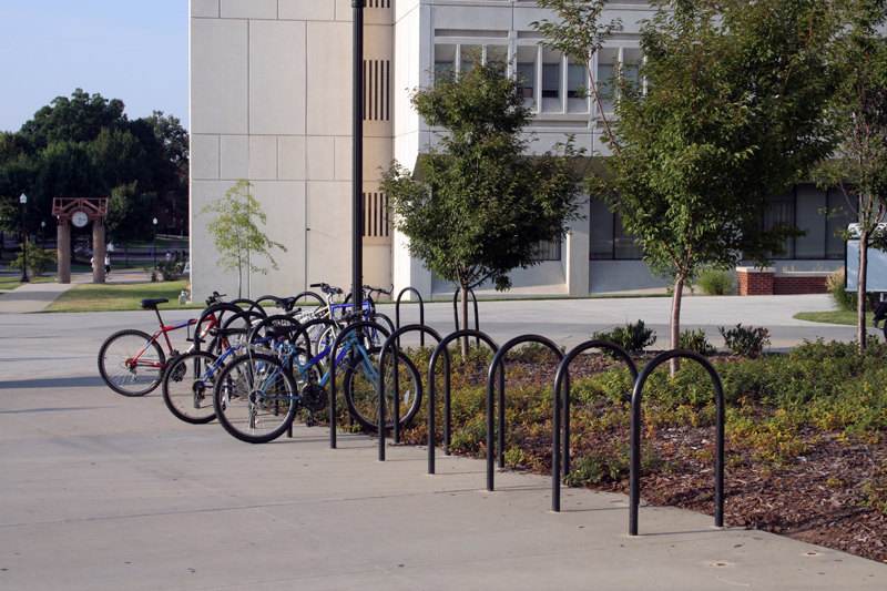 Black U-racks are installed in an L shape around a bush in front of a school, several bikes locked to them