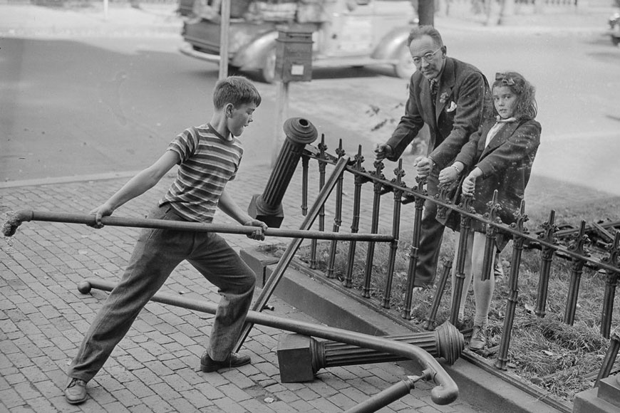 A boy with a large pipe breaks a fence held by an older man and a girl; bollards and handrails lie on the ground