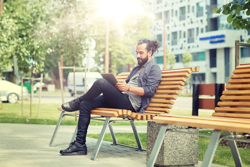 Man sits on urban park bench reading tablet, office buildings and trees behind him