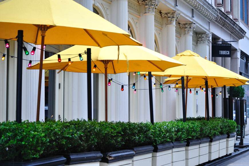 Yellow patio umbrellas are strung with colorful outdoor lights behind a low wall made of planters