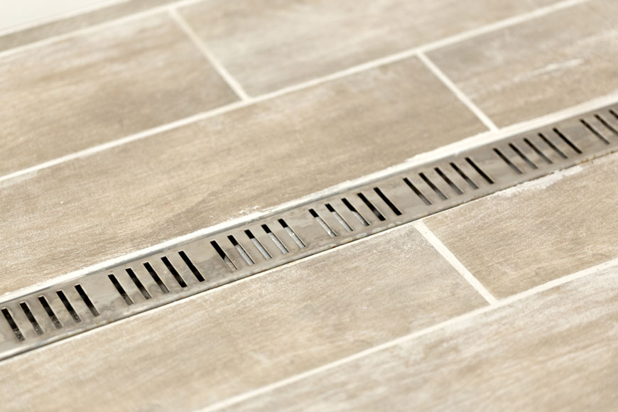 A thin stainless steel trench grate drains water inside a tiled shower